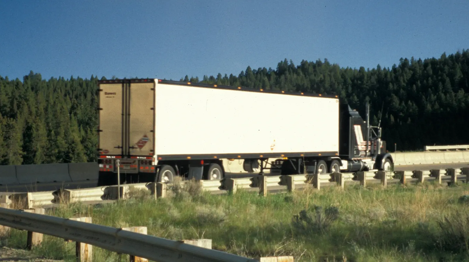 A tractor pulling a white dry van trailer along a highway with evergreen forest behind